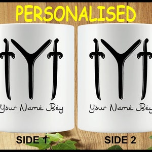 May include: White ceramic mug featuring a black design of a symbol with two swords on either side. The text "Your Name Bey" is printed on the mug.
