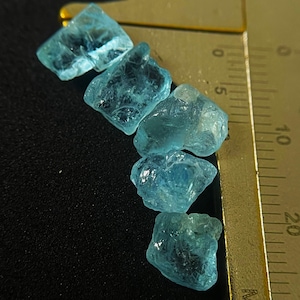 May include: Five rough, light blue gemstones are arranged diagonally on a black surface next to a ruler. The gemstones have a crystalline appearance and varying shapes. The ruler shows measurements in centimetres.