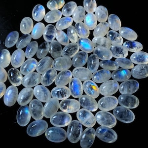 Op de afbeelding: Een verzameling ovaalvormige maansteen edelstenen. De stenen zijn doorschijnend met een melkachtige witte basis en vertonen een blauwe adularescentie. De edelstenen liggen op een zwarte ondergrond, wat hun iriserende kwaliteit benadrukt.