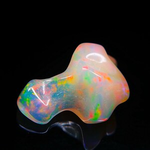 Könnte beinhalten: Ein polierter Opal-Edelstein mit einer einzigartigen, organischen Form. Der Stein zeigt ein Farbspiel mit Blitzen von Blau, Grün, Orange und Rot auf einer milchig-weißen Basis. Der Edelstein ist vor einem schwarzen Hintergrund platziert.