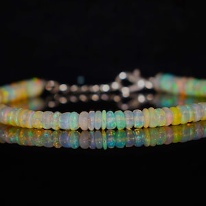Puede incluir: Una pulsera compuesta por pequeñas cuentas de ópalo redondas y translúcidas que muestran destellos iridiscentes de verde, naranja y amarillo. La pieza cuenta con un cierre de mosquetón metálico y una cadena de extensión ajustable.