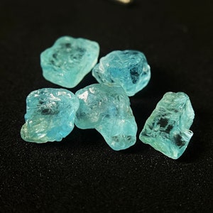 May include: Five rough aquamarine gemstones in a light blue hue. The stones have a crystalline appearance and irregular shapes, with a slightly translucent quality. They are displayed against a dark, textured background.