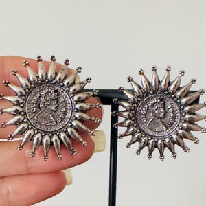May include: Silver-toned stud earrings with a coin design at the center, surrounded by a sunburst pattern. The earrings are round and feature a detailed profile on the coin. The earrings are displayed on a black stand.