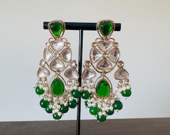 May include: Gold-toned chandelier earrings with green and clear gemstones and white pearl accents. The earrings feature a geometric design with a large green teardrop-shaped stone at the bottom and a smaller green stone at the top.