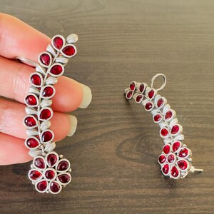 May include: Silver-toned earrings with a floral and leaf design, adorned with vibrant red gemstones. The earrings curve upwards, with a cluster of gemstones at the base and along the stem.
