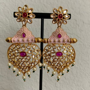 May include: Gold-toned, ornate earrings with a floral top and heart-shaped bottom. The earrings feature clear and ruby-colored stones, with a pink enamel accent. Small pearls and green beads dangle from the bottom, creating an elegant, eye-catching design.