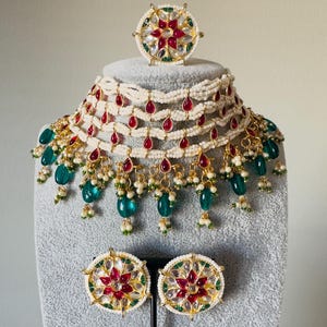 May include: An ornate jewelry set featuring a necklace, ring, and earrings. The necklace has multiple strands of white beads with red teardrop-shaped accents and green gemstone dangles. The ring and earrings have a floral design with red and white accents.