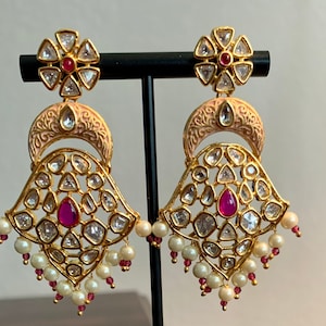 May include: A pair of gold earrings with pink and white stones. The earrings have a teardrop shape with a smaller flower-shaped piece at the top. The earrings are adorned with pearls and pink stones.