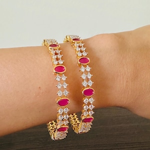 May include: Two gold-toned bangles with alternating oval magenta gemstones and clear, sparkling crystals. The bracelets are worn on a wrist, showcasing the intricate design and vibrant colors of the jewelry.