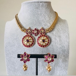 May include: A gold-toned necklace and earring set. The necklace features a chain with a pendant of two large, round, red and white stone-studded medallions. The earrings are flower-shaped with red and white stones and a gold-toned bead.