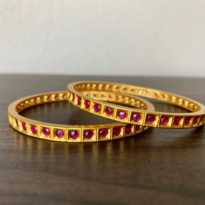 May include: Two gold bangles with a row of pink gemstones set in square bezels.