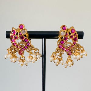 May include: A pair of gold-toned stud earrings with a peacock design. The earrings feature ruby-colored and clear gemstones, with dangling pearl accents. The earrings are displayed on a black stand against a white background.
