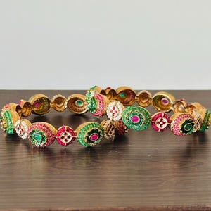 May include: Two gold bangles with green and pink stones and white pearls. The bangles are stacked on top of each other.
