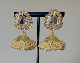 May include: A pair of gold earrings with a teardrop-shaped design. The earrings have a clear glass stone in the center and are surrounded by smaller clear stones. The bottom of the earrings have a gold ball with a fringe of white pearls.