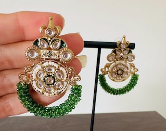 May include: Gold-toned earrings with a detailed design. The earrings feature clear, round accents, white and green enamel, and a green beaded hoop. One earring is held by a hand, and the other is displayed on a black stand.