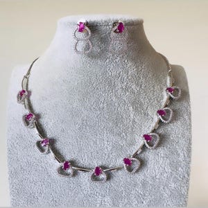 May include: A silver-toned necklace and matching earring set featuring oval-cut vibrant pink stones. The necklace consists of linked segments, each holding a pink stone surrounded by small clear crystals. The earrings mirror this design.