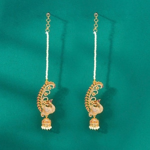 May include: Two gold-colored, ornate earrings with a peacock design. Each earring features a dangling pearl strand, a peacock-shaped pendant with red and clear gemstones, and a bell-shaped bottom with small pearls. The background is a teal green.