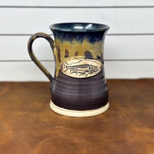 May include: A ceramic mug with a dark purple base and a tan, drippy glaze near the rim. The mug features a centered, oval-shaped relief medallion depicting a detailed fish. The handle is curved and matches the dark purple glaze of the body.