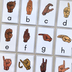 May include: A set of 12 flashcards featuring the American Sign Language alphabet from A to L. Each card has a white background with a black border and a brown hand sign representing a letter. The letter is printed in black below the hand sign.
