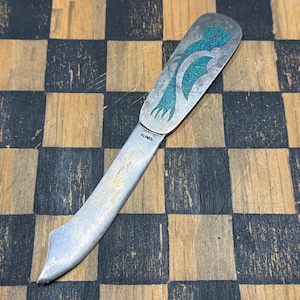May include: A silver-toned metal letter opener featuring a flat handle inlaid with a textured teal stone design of a bird. The blade is smooth and metallic with the word ALPAC stamped near the base of the handle, showing a curved edge.