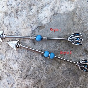 May include: Two silver arrow-shaped body jewellery pieces with blue beads. The top piece has a feather design on the end. The bottom piece has a simple arrowhead on the end. The text "Style 1" and "Style 2" are written on the image.