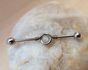 May include: A silver industrial barbell earring with a central clear stone and two silver balls on each end. The center stone is held in place by a silver wire loop and wrapped with copper wire.