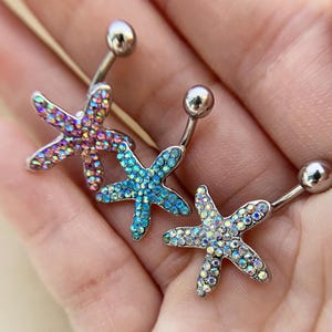 May include: Three belly button rings featuring starfish pendants. The starfish are adorned with iridescent rhinestones in shades of pink, purple, blue, and clear. Each ring has a curved bar with a small ball at each end.