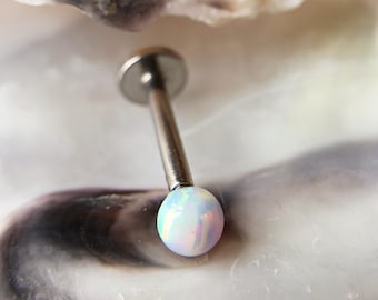 Puede incluir: Primer plano de un piercing labret plateado con una bola redonda iridiscente similar al ópalo. El piercing tiene un disco plano en un extremo y un poste largo y delgado. La bola de ópalo refleja tonos blancos, azules y rosas.