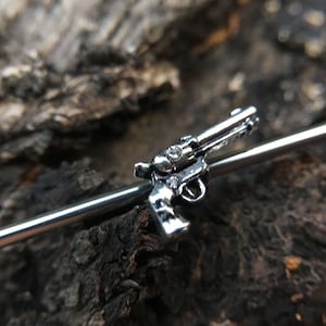 May include: A silver-toned gun charm attached to a silver-toned metal bar. The charm is a detailed miniature replica of a revolver.