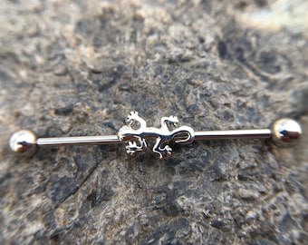May include: Silver barbell with a lizard charm in the center. The barbell has two silver balls on either end.