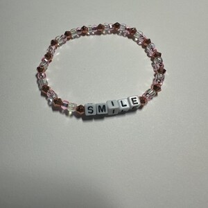 Op de afbeelding: Een roze en gouden kralen armband met het woord "SMILE" gespeld in witte letter kralen.