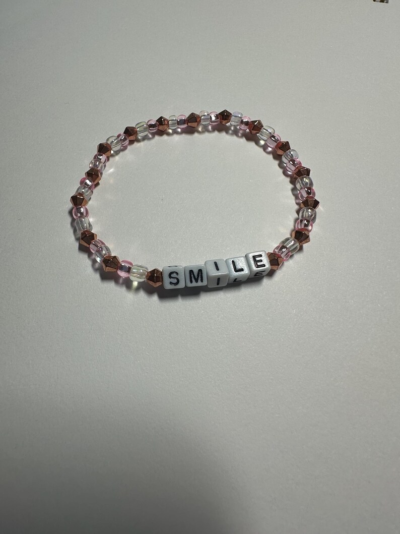Op de afbeelding: Een roze en gouden kralen armband met het woord "SMILE" gespeld in witte letter kralen.