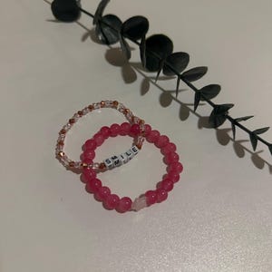 Op de afbeelding: Twee armbanden, een roze met witte kralen die "SMILE" spellen en een heldere met koperen kralen.