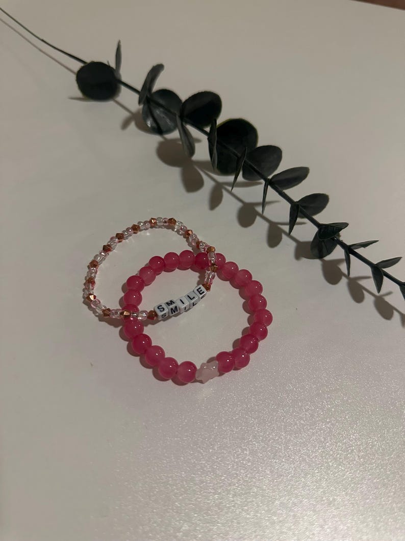 Op de afbeelding: Twee armbanden, een roze met witte kralen die "SMILE" spellen en een heldere met koperen kralen.