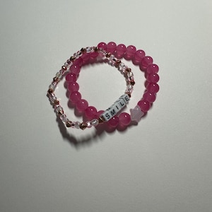 Op de afbeelding: Twee roze kralenarmbanden, een met een sterbedel en de andere met het woord "SMILE" gespeld in witte letters.