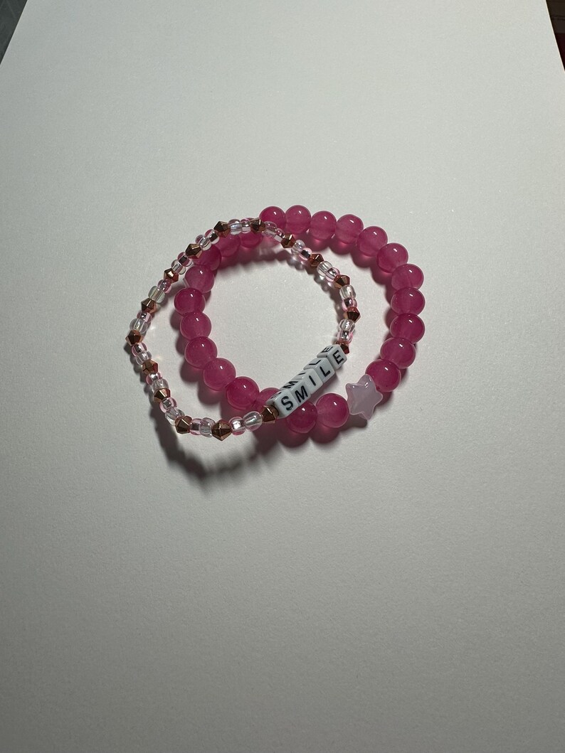 Op de afbeelding: Twee roze kralenarmbanden, een met een sterbedel en de andere met het woord "SMILE" gespeld in witte letters.