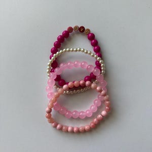 Może przedstawiać: Cztery bransoletki ułożone jedna na drugiej. G&oacute;rna bransoletka jest r&oacute;żowa z złotymi literami tworzącymi napis "LOVE". Następna bransoletka jest biała z małymi okrągłymi koralikami. Trzecia bransoletka jest jasnor&oacute;żowa z większymi okrągłymi koralikami. Dolna bransoletka jest ciemnor&oacute;żowa z mniejszymi okrągłymi koralikami.