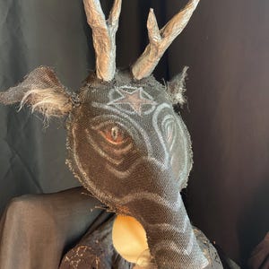 May include: A handcrafted mask with a deer-like design. The mask features a dark, textured fabric with painted details, including a star and eye design. Silver-colored antlers and feathery accents complete the look. The mask is likely for costume or decorative purposes.
