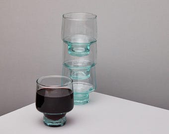 May include: A set of four clear glass tumblers stacked on top of each other. The tumblers have a light blue tint and a slightly textured surface. One tumbler is filled with red wine.