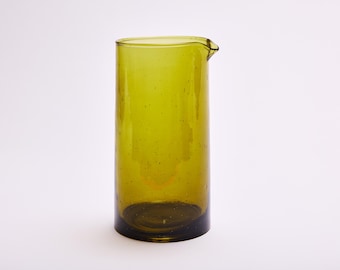 May include: A tall, olive-green glass pitcher with a spout. The cylindrical body has a dark base and a slightly flared rim. The glass has small bubbles throughout, giving it a textured appearance. The pitcher is empty and sits against a white background.