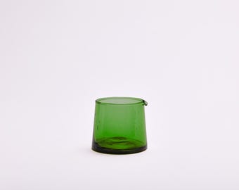 May include: A small, translucent green glass creamer or pitcher with a wide, tapered body and a subtle pouring spout. The glass features a smooth, rounded rim and a flat base, showcasing a vibrant emerald hue and a simple, functional design.