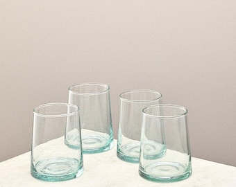 May include: Four clear, cylindrical drinking glasses with a slightly flared shape. The glasses are made of transparent glass, with a subtle blue-green tint at the base. They are arranged on a white surface against a neutral background.
