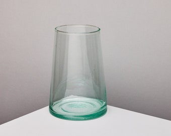May include: A clear glass vase with a slightly flared shape. The vase has a wide base and a narrow opening. The glass has a slight green tint. The vase is empty and sits on a white surface.