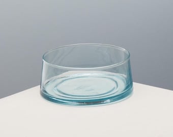 May include: A clear, round, shallow glass bowl with a light blue tint. The bowl sits on a white surface against a gray background. The bowl's design is simple and modern, with a slightly angled rim.