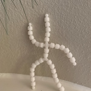 May include: A white articulated figure made of small, round beads. The figure is posed in a standing position with arms raised and legs spread.