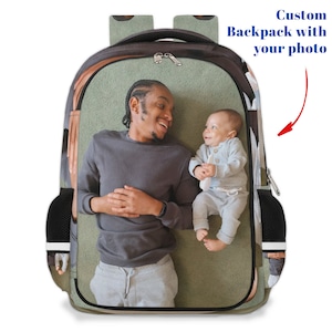 May include: A personalised backpack featuring a photo of a man and a baby boy lying on a green blanket. The backpack has a black mesh pocket on the side and a zipper closure.
