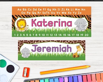 May include: Classroom name labels with a jungle safari theme. The labels feature the names "Katerina" and "Jeremiah" in pink and purple, with animal illustrations. The top label has a lion and elephant, while the bottom has an elephant and giraffe. The labels are on a white background.