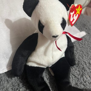 Könnte beinhalten: Ein weiß-schwarzer Panda Beanie Baby mit einem roten Band um den Hals. Der Beanie Baby hat ein Ty-Etikett an seinem Ohr, auf dem "Beanie Babies Baby" steht.