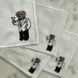 May include: White square cocktail napkins with an embroidered brown bear wearing a tuxedo and holding a martini glass. The bear is the focal point of the design, repeated on multiple napkins.