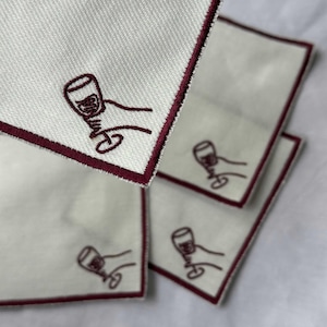 Embroidered white cotton cocktail coasters with burgundy stitching of an embroidered hand holding a wine glass